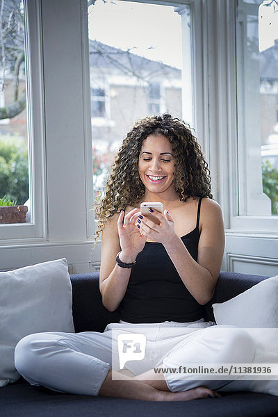 Lächelnde Frau sitzt in der Nähe des Fensters und schreibt eine SMS auf ihrem Handy