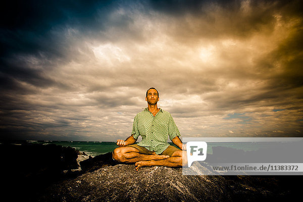 Mann meditiert auf einem Felsen am Meer unter Wolken