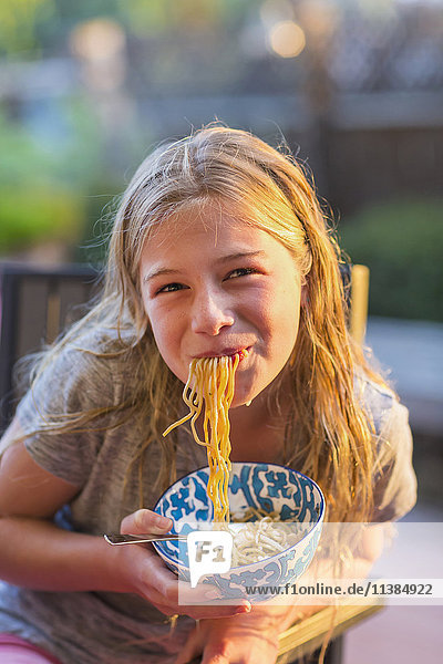 Noodles hanging from mouth of Caucasian girl holding bowl