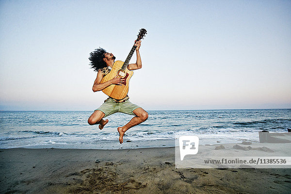Mixed Race Mann spielt Gitarre und springt am Strand