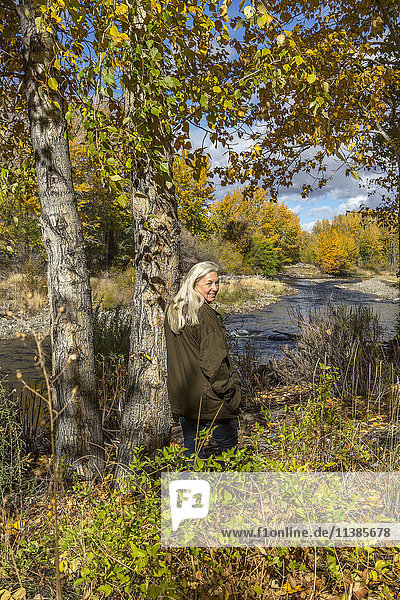 Lächelnde kaukasische Frau lehnt an einem Baum in der Nähe eines Flusses