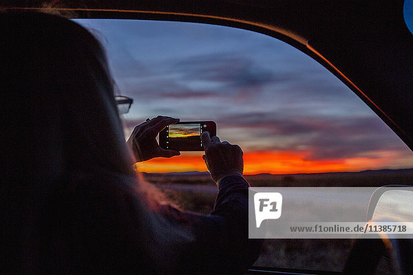 Kaukasische Frau im Auto fotografiert Sonnenuntergang mit Handy