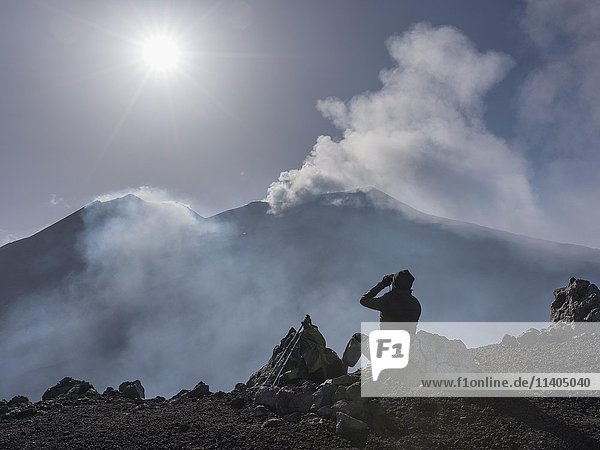 Bergwanderer schaut auf zwei rauchende Ostkrater im Gegenlicht  Blick vom Aussichtspunkt Rocca della Valle  Ätna  Vulkan  Nordostflanke  Sizilien  Italien  Europa
