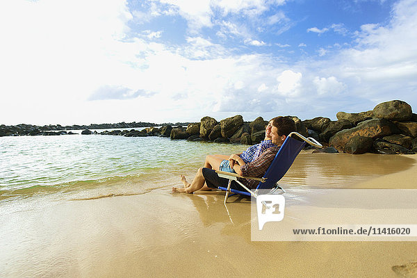 'A couple sits in a beach chair together facing the ocean on Lydgate Beach; Kauai  Hawaii  United States of America'