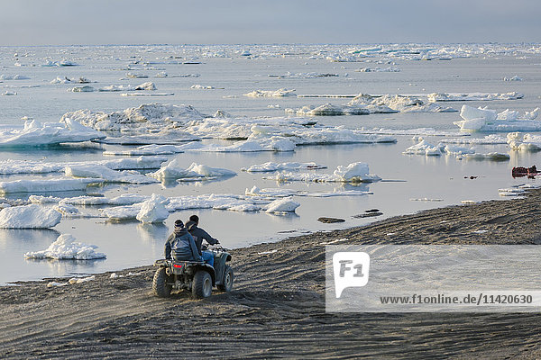 Zwei einheimische Männer aus Alaska fahren mit einem ATV einen Sandstrand entlang des mit Meereis gefüllten Arktischen Ozeans im Sommer hinunter  North Slope  Arktisches Alaska  USA