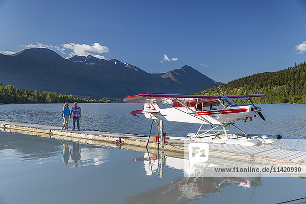Eine Frau und ein Mann stehen auf einem Dock  an dem ein Wasserflugzeug angedockt ist  Trail Lake Float Plane Base  Moose Pass  Kenai Peninsula  Southcentral Alaska  USA