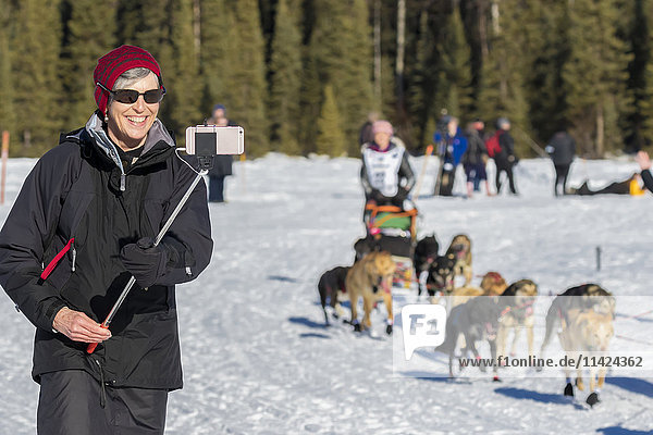 A woman tourist takes an smart phone photo of a musher in the start of the Iditarod Race on Willow Lake  Alaska  USA  Winter.