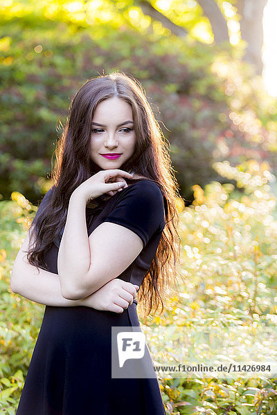 Ein junges Teenager-Mädchen in einem modischen schwarzen Kleid steht in einer nachdenklichen Pose in einem sonnigen Park; Vancouver  British Columbia  Kanada'.