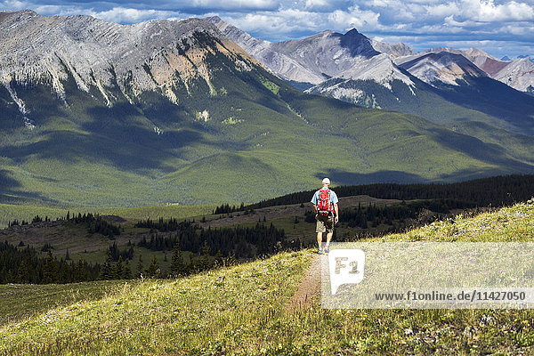 'Male hiker walking along a hillside trail overlooking mountain range and valley with a cloudy sky  West of Bragg Creek; Alberta  Canada'