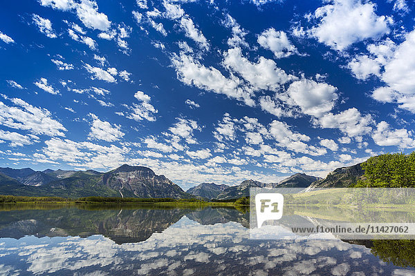 Dramatische Wolken  die sich auf einem ruhigen Bergsee mit einer Bergkette in der Ferne und blauem Himmel spiegeln; Waterton  Alberta  Kanada'.