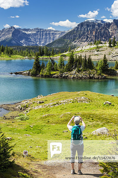 Wanderin  die auf einem Wanderweg steht und mit ihrem Handy ein Foto von einem Alpensee mit Bergen  blauem Himmel und Wolken macht; Banff  Alberta  Kanada'.
