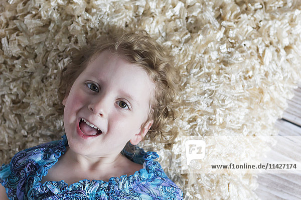 Ein junges Mädchen mit lockigem blondem Haar liegt auf dem Boden auf einem weichen Teppich und schaut spielerisch in die Kamera und lacht; St. Albert  Alberta  Kanada'