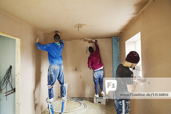 Drei Männer  zwei Stuckateure mit Stelzen  die hoch oben an den Wänden eines im Bau befindlichen Hauses frischen Putz glätten.