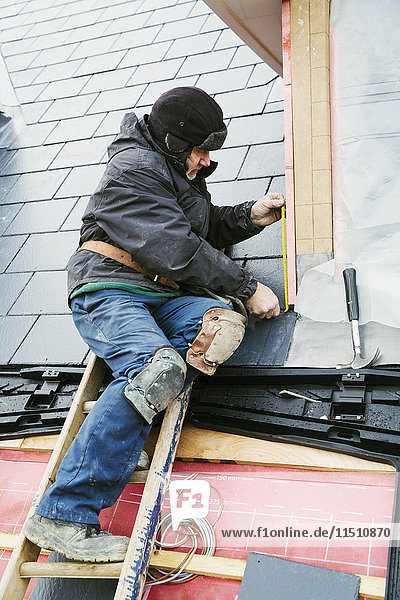 Ein Mann an der Spitze einer Leiter auf einem Hausdach  der Ziegel auf einem Gaubendach befestigt.