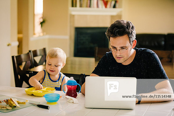 Vater und kleiner Sohn sitzen am Tisch  Sohn isst  Vater benutzt Laptop