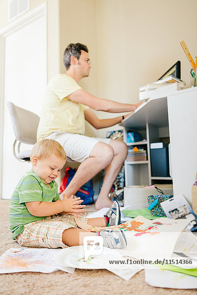 Father using computer whilst young son plays on floor