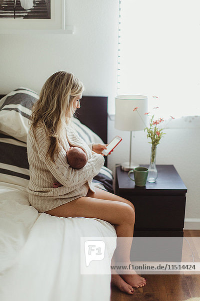 Mid adult woman sitting on bed looking at smartphone whilst cradling new born baby daughter