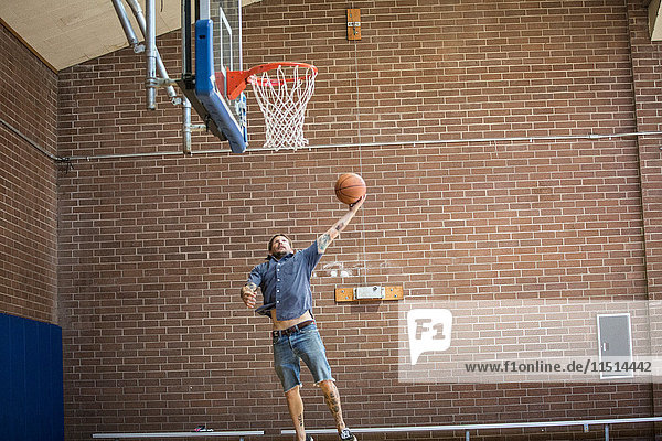 Tätowierter Mann springt und zielt mit dem Ball auf das Basketballnetz auf dem Platz