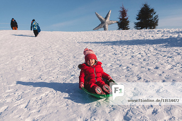 Mädchen mit roter Strickmütze rodelt einen schneebedeckten Hügel hinunter