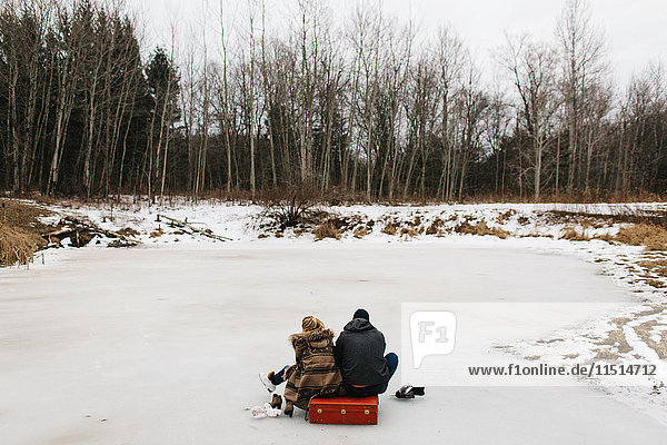 Couple sitting on red suitcase in middle of frozen lake  Whitby  Ontario  Canada