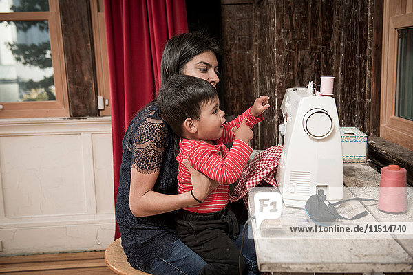 Männliches Kleinkind auf dem Schoß der Mutter an der Nähmaschine sitzend