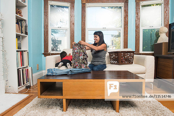 Männliches Kleinkind klettert auf Sofa  während die Mutter Wäsche faltet