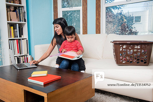 Frau benutzt Laptop  während der Kleinkind-Sohn das Notebook auf dem Sofa betrachtet