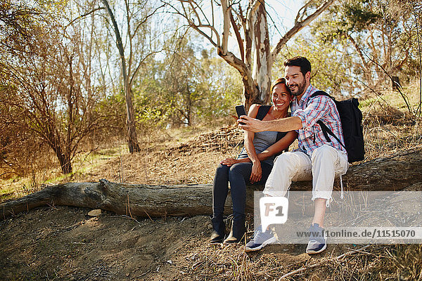 Mid adult couple hiking  sitting on log taking smartphone selfie