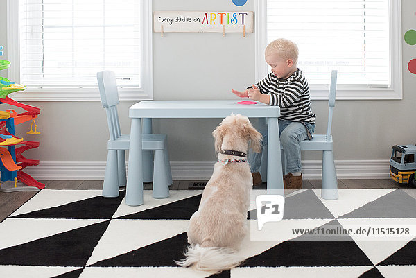 Junge spielt mit rosa Teig auf dem Tisch  Hund sitzt neben dem Tisch und schaut zu.