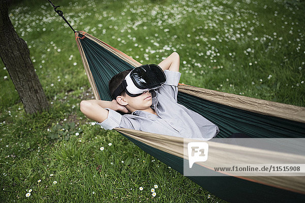 Gemischtrassiger Junge liegt in der Hängematte und trägt eine Virtual-Reality-Brille