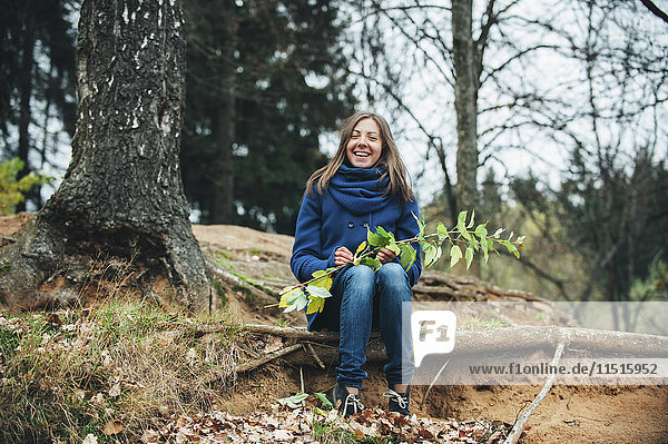 Lächelnde kaukasische Frau sitzt auf einer Baumwurzel und hält einen Ast