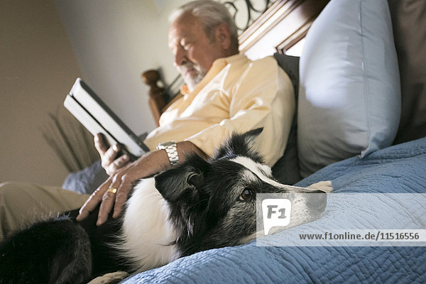 Older man laying on bed with dog reading digital tablet