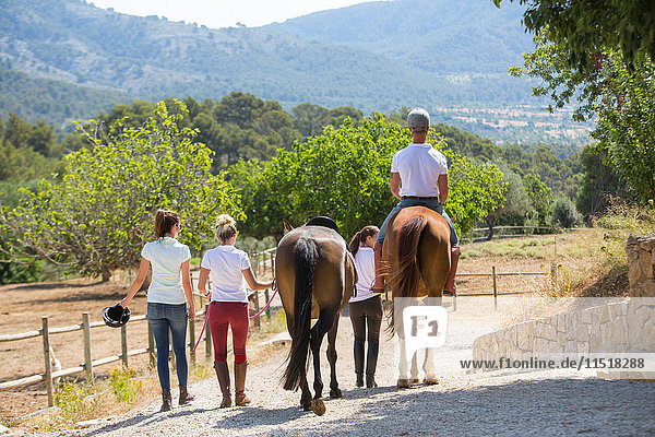Rear view of female groom leading horse rider along paddock at rural stables