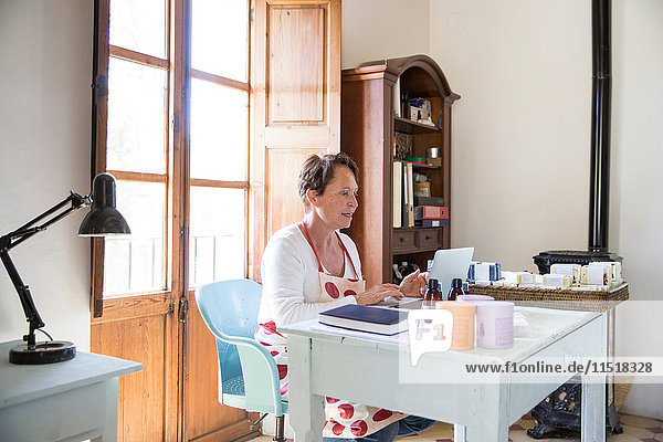 Reife Frau tippt auf Büro-Laptop in handgemachter Seifenwerkstatt