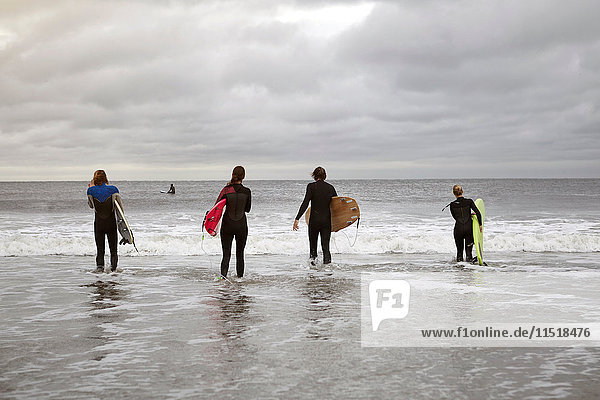 Rear view of four young adult surfers carrying surfboards into sea on Rockaway Beach  New York  USA