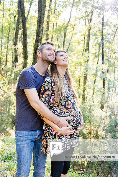 Schwangeres Paar im Wald umarmt sich  schaut lächelnd auf