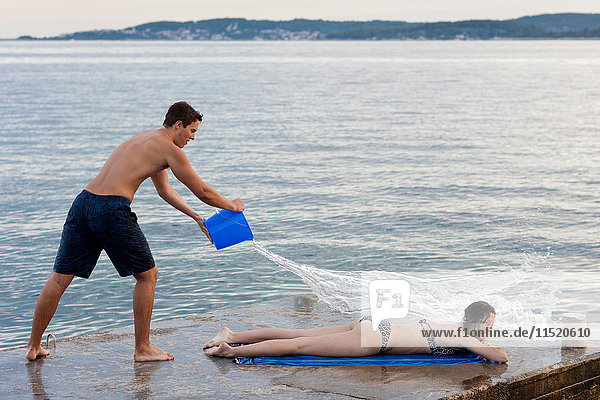 Junger Mann wirft einen Eimer Wasser über einen weiblichen Sonnenbadenden  Orebic  Kroatien