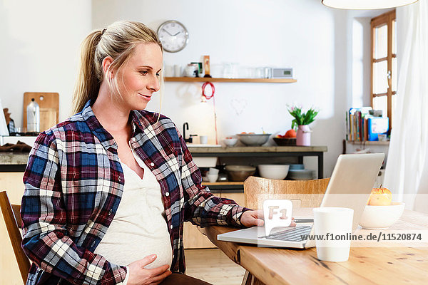 Schwangere Frau hält Bauch  während sie auf den Laptop auf dem Tisch schaut