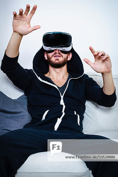 Mittelgroßer erwachsener Mann auf Sofa mit erhobenen Armen  der ein Virtual-Reality-Headset betrachtet