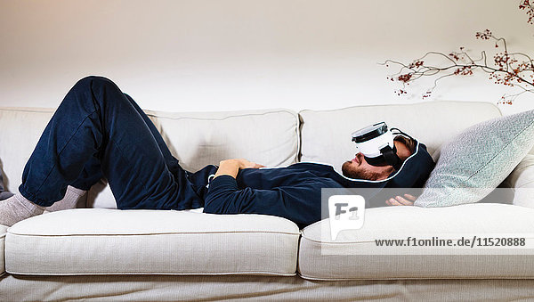 Mittelgroßer erwachsener Mann  der auf dem Sofa liegt und sich ein Virtual-Reality-Headset anschaut