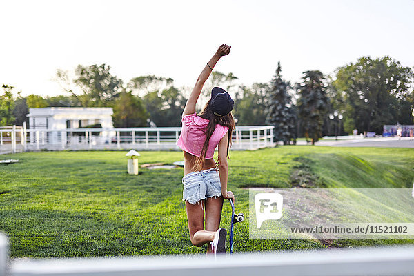 Junge Frau geht durch den Park  trägt Skateboard  schlägt Luft  Rückansicht
