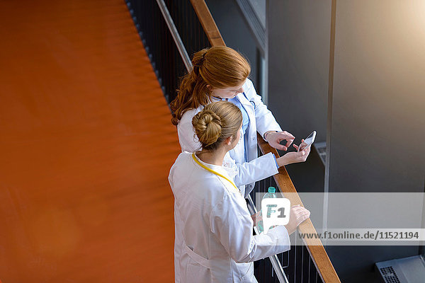 Hochwinkelansicht von zwei Ärztinnen auf dem Krankenhausbalkon mit Blick auf das Smartphone
