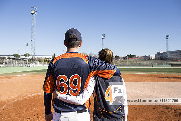 Rückansicht eines männlichen und weiblichen Baseballspielers  der sich auf einem Baseballfeld umarmt.