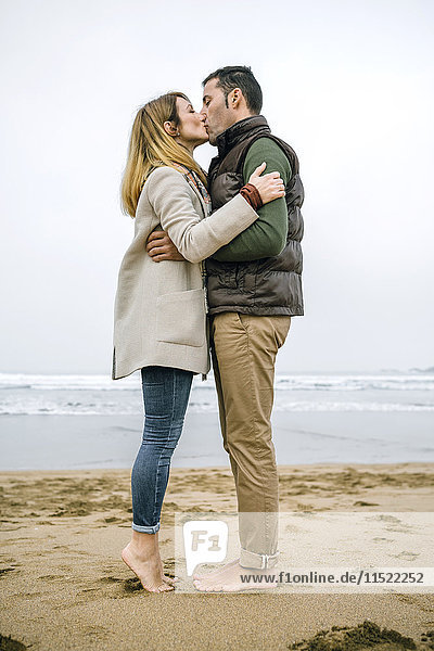 Ein Paar steht barfuß am Strand und küsst sich.