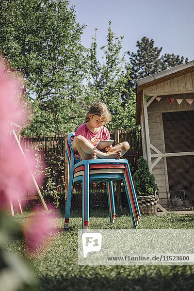 Mädchen sitzend auf Stuhlstapel im Garten mit Tablette