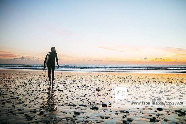 Junge Frau mit Surfbrett  die bei Sonnenuntergang am Strand spazieren geht.