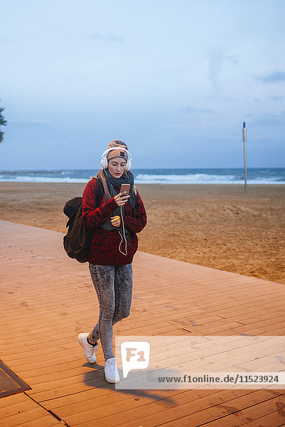 Junge Frau hört Musik auf einem Smartphone  während sie in der Abenddämmerung am Strand spazieren geht.