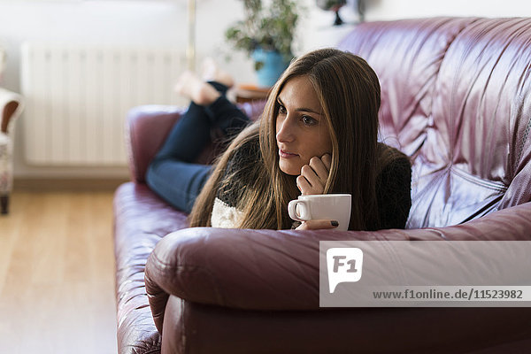 Junge Frau liegt auf der Couch und hält eine Tasse Kaffee.