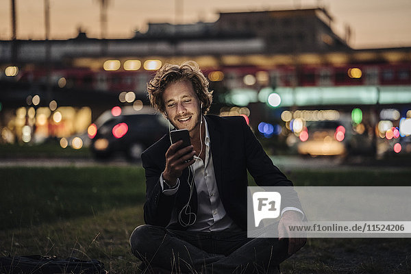 Lächelnder Geschäftsmann auf der Wiese in der Abenddämmerung mit Handy und Kopfhörer