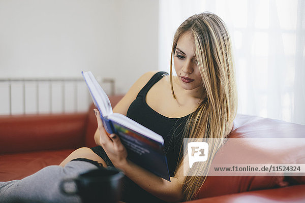 Junge Frau sitzt auf der Couch und liest ein Buch.